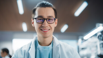 Obraz premium Portrait of an Asian Scientist Posing for Camera, Smiling. Young Male Biologist in a White Coat Working in a Modern Laboratory, Researching Medical Drug Products and Genome Editing