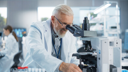 Medical Research and Development Laboratory: Senior Caucasian Male Scientist in Glasses Looking at a Sample in a Petri Dish Under a Microscope in an Advanced Biotechnology Lab