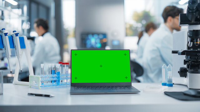 Laptop Computer with Green Screen Mock Up Display Standing on a Table in a Science Laboratory Next to a Microscope and Test Tubes with Biological Samples. Anonymous People Working in the Background