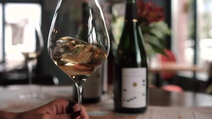 A close-up shot of an elegant hand swirling a glass of white wine, showcasing the wine's clarity and texture. In the background, out-of-focus wine bottles and a softly lit restaurant