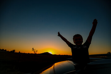 Silhouette of child reaching out sunroof of vehicle with arms up on country road at sunset