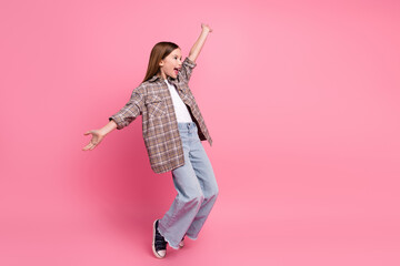 Young cheerful girl in trendy plaid shirt posing against a pink background with outstretched arms.