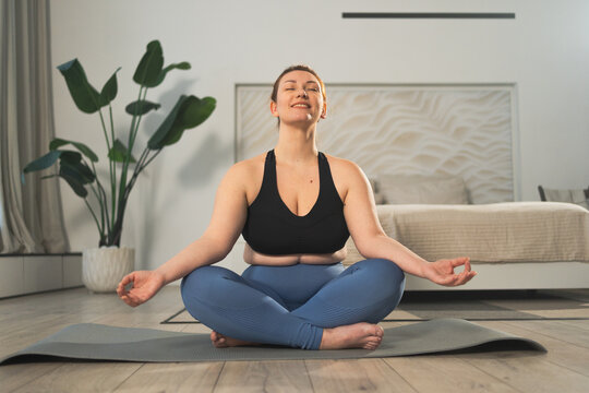 Yoga mindfulness meditation. Woman practicing yoga in living room at home. Plus size female sitting in lotus pose meditating smiling relaxing indoor. Girl doing breathing practice. Yoga at home