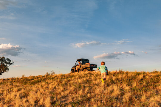 Little boy running up hill towards Dad sitting in off-road vehicle on farm