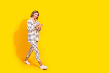 Confident young woman in a stylish checkered suit walking with a tablet against a vibrant yellow background.