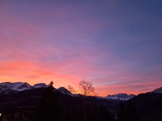 Sunset over a mountainous landscape. The sky is vibrant with shades of orange, pink, and purple. Sun sets behind the majestic snowy peaks