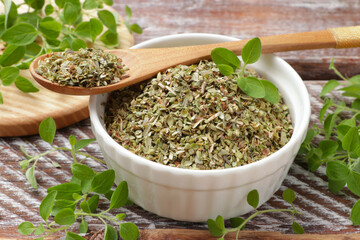 Dried oregano in bowl, spoon and green leaves on wooden table, closeup