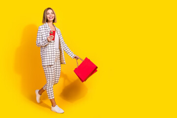 Confident young businesswoman in fashionable suit holding phone and red shopping bags against vibrant yellow background
