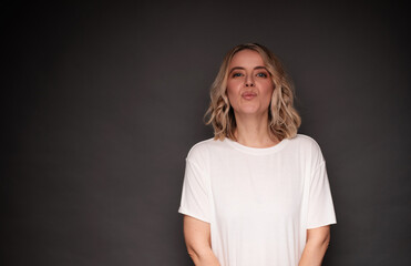 A cheerful blonde woman in a white t-shirt poses against a gray background, blowing a kiss with a playful expression, perfect for themes of affection, fun, and positive emotions.