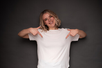 A cheerful woman wearing a white t-shirt points at herself with both hands, smiling brightly. The photo is set against a dark background, ideal for themes of self-confidence and positivity.