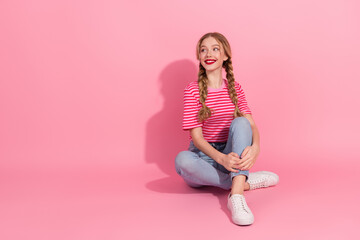 Charming young woman with braids sits against a pink background, smiling brightly in a striped top and jeans.