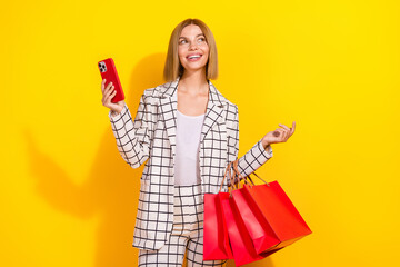 Charming young woman holding shopping bags and smartphone against a vibrant yellow background showcasing her suit and beautiful smile