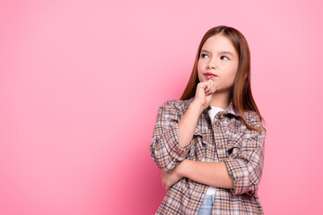 Thoughtful young girl in casual checkered outfit posing against pink background with curious...