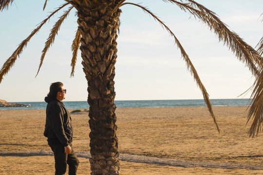 A middle-aged man looks out at the horizon next to a palm tree at sunset, on Comarruga beach, Tarragona