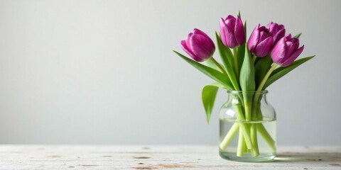 A simple arrangement of purple tulips in a clear glass vase on a rustic white table against a neutral backdrop