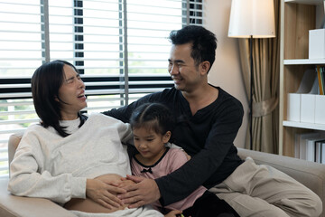Fototapeta premium A pregnant woman and her two children are sitting on a couch. The woman is wearing a white shirt and the children are wearing pink. Scene is happy and loving 