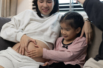 A pregnant woman and her two children are sitting on a couch. The woman is wearing a white shirt and the children are wearing pink. Scene is happy and loving
