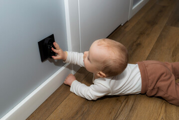 A seven-month-old Caucasian baby touches a black electrical outlet on the wall.