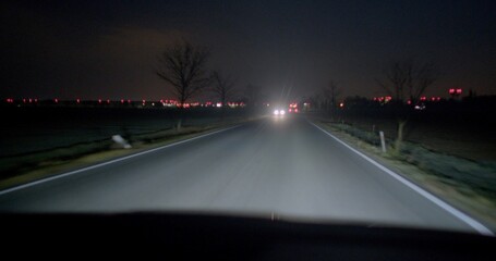 A car is driving down a winding road at night, surrounded by shadows and the glow of streetlights illuminating the path ahead
