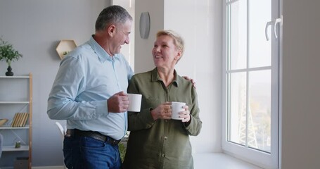 Happy retired senior couple spending time at home. Loving husband brings coffee to wife who is looking out window in living room. Man joins woman, they drink coffee, admire view, discuss neighborhood