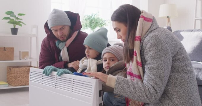 Worried frozen young family with children wearing winter outwear sitting on sofa in chilly living room at home warming up hands on electric heater. Heating problems concept. 4k video.