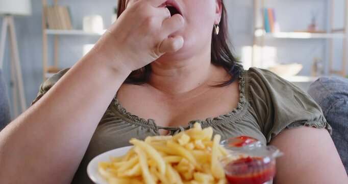 Closeup of young fat overweight woman sitting in living room at home and eating tasty french fries with ketchup. Plus size plump girl enjoying unhealthy fast food. Nutrition concept. 4k video.