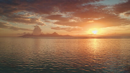Fototapeta premium Drone fly over sunset sand beach on Tahiti tropical island. Bright orange evening sky reflects on calm ocean water waves. Beautiful sunrise landscape. Remote wild nature paradise, exotic summer travel