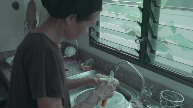 An elderly woman skillfully washes vegetables in a sunlit kitchen, showcasing a moment of everyday life. The bright surroundings highlight the engaging process of preparing fresh ingredients for a mea