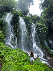 A women stands in front of a lush, green waterfall surrounded by dense foliage. The cascading water...