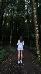 A girl stands on a forest path surrounded by tall trees and dense foliage, gazing into the lush greenery. The scene captures the serene and immersive experience of being in nature.