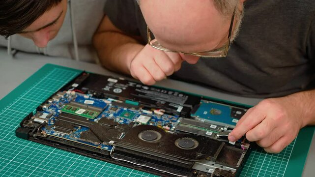 Father with son analyzing laptop components, considering repair options. Male and boy closely inspecting notebook internals, debating fixes. Guy and kid examining computer parts, brainstorming