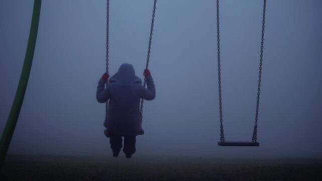 Woman sits on children swings in eerie dark foggy playground