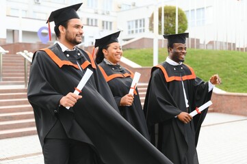Group of happy smiling multicultural people in graduation gowns and caps with diplomas in hands graduate university outdoors in campus looking at camera. Graduation from college, university concept.
