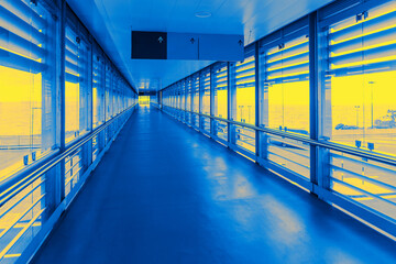 Modern airport walkway with large glass windows and metal railings, bathed in striking blue and yellow lighting, creating a futuristic and vibrant atmosphere