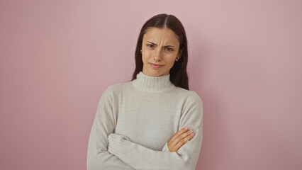 Fototapeta premium Attractive hispanic woman standing with arms crossed in front of a pink wall, wearing a beige sweater, showcasing a skeptical expression