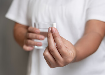 Woman in white t-shirt holding white pill in fingers and water glass in hand closeup