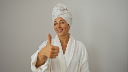 Mature woman with brunette hair giving a thumbs-up in a white robe and towel turban on an isolated white background