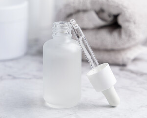 Opened white glass dropper bottle with pipette on marble near cosmetics and towel close up