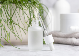 Opened white dropper bottle with pipette on marble near cosmetics and green plant close up