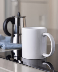 White coffee mug on induction stove against moka pot on blue dish cloth closeup, mockup