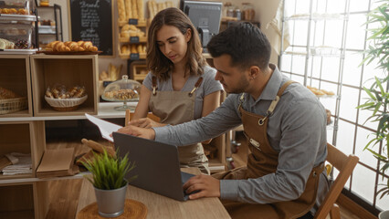 Man and woman working together in a bakery interior, wearing aprons and using a laptop with pastries displayed in the background, indicating a collaborative work environment.