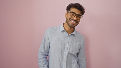 Young man with glasses and beard smiling confidently against a stylish pink background wearing...