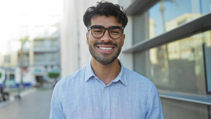Obraz premium Handsome young hispanic man with a beard smiling outdoors on a city street wearing glasses and a striped shirt in an urban environment on a sunny day.