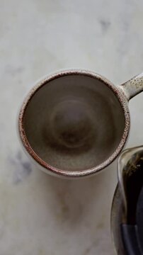close-up vertical scene of pouring coffee from the filter coffee maker to a round scandic mug on a marble kitchen table. moody natural light cozy scene