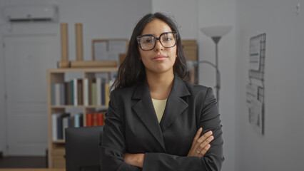 Confident woman with brunette hair, arms crossed, wearing glasses, standing in an indoor office setting.