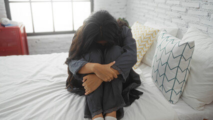 Woman sitting alone in a bedroom, looking distressed, with long brunette hair, wearing dark clothes, and hugging her knees on a white bed with decorative pillows.