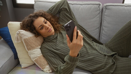 Woman relaxing indoors on mobile phone in cozy apartment with brunette curls and casual clothes, comfortably reclining on a sofa surrounded by cushions.