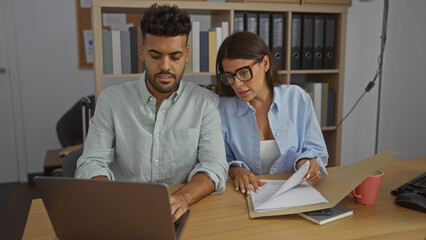 Man and woman working together in an office setting with documents and laptop, indicating a collaborative business environment