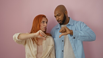 Interracial couple showing thumbs down while standing against a pink wall, conveying disagreement and frustration in their relationship