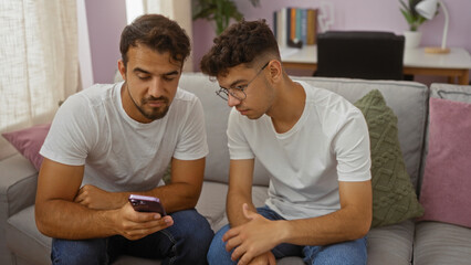 Hispanic father and son bonding over a mobile phone on a cozy couch in a living room, showcasing family love and togetherness in an indoor setting.